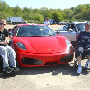 Two people in wheelchairs posing beside a red Ferrari at an outdoor car event.