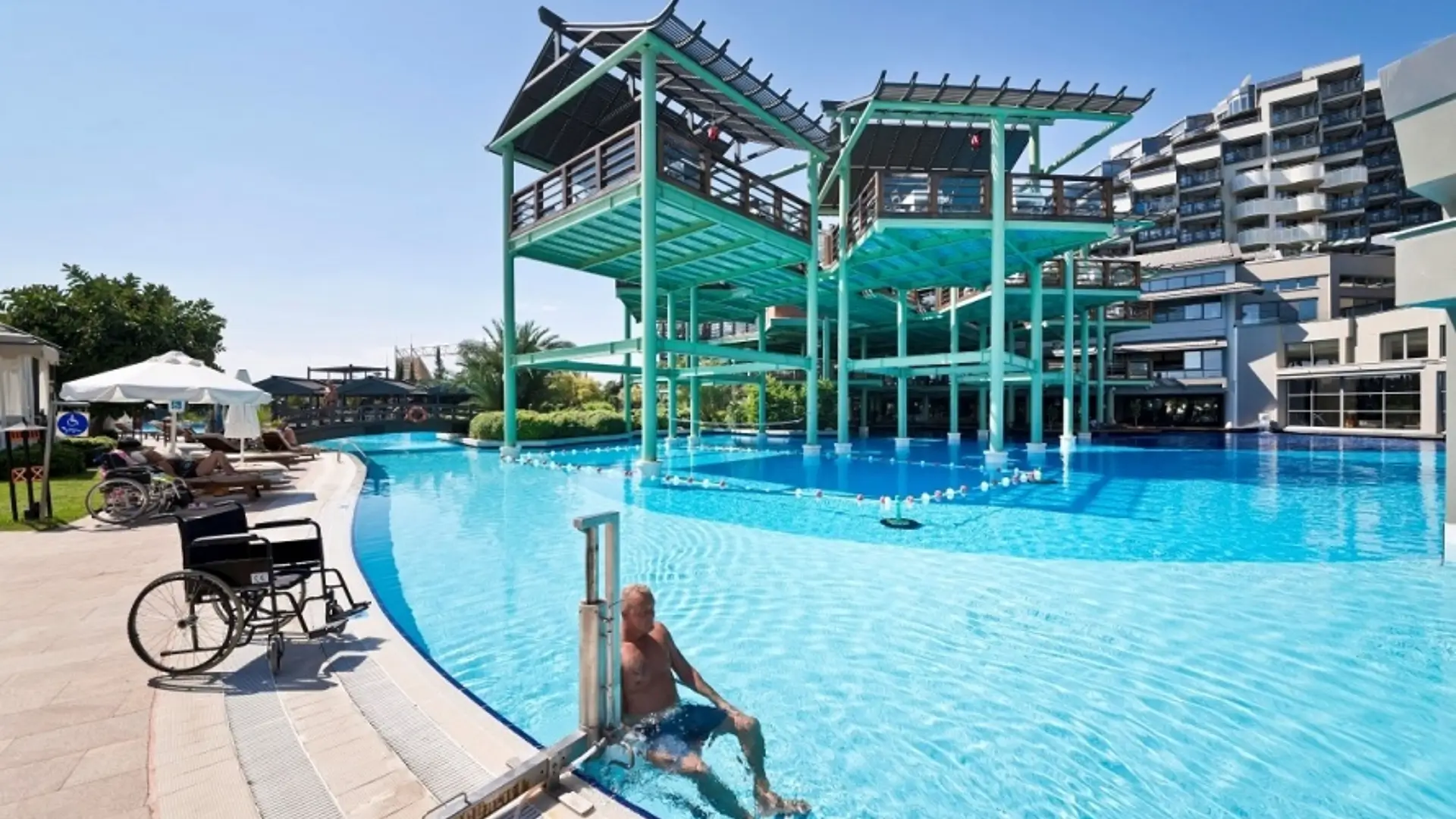 Man using pool hoist beside wheelchairs at resort.