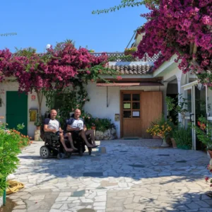 Two men in wheelchairs outside flower-covered villa.
