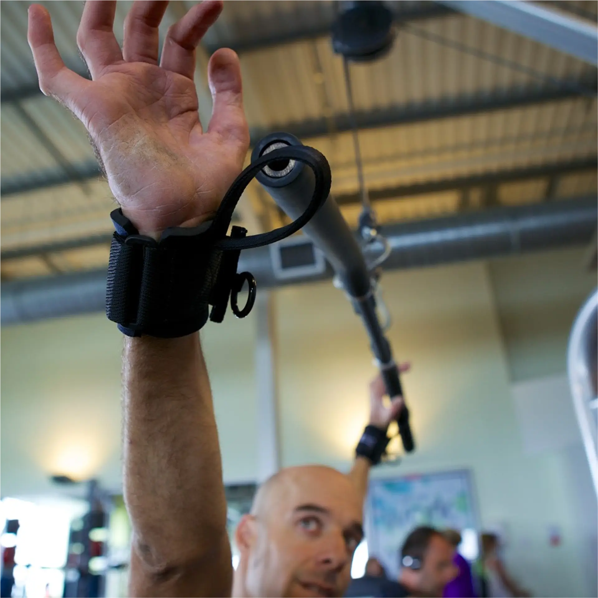 Close-up of a wrist strap gripping a gym cable handle, arm raised overhead in a gym.