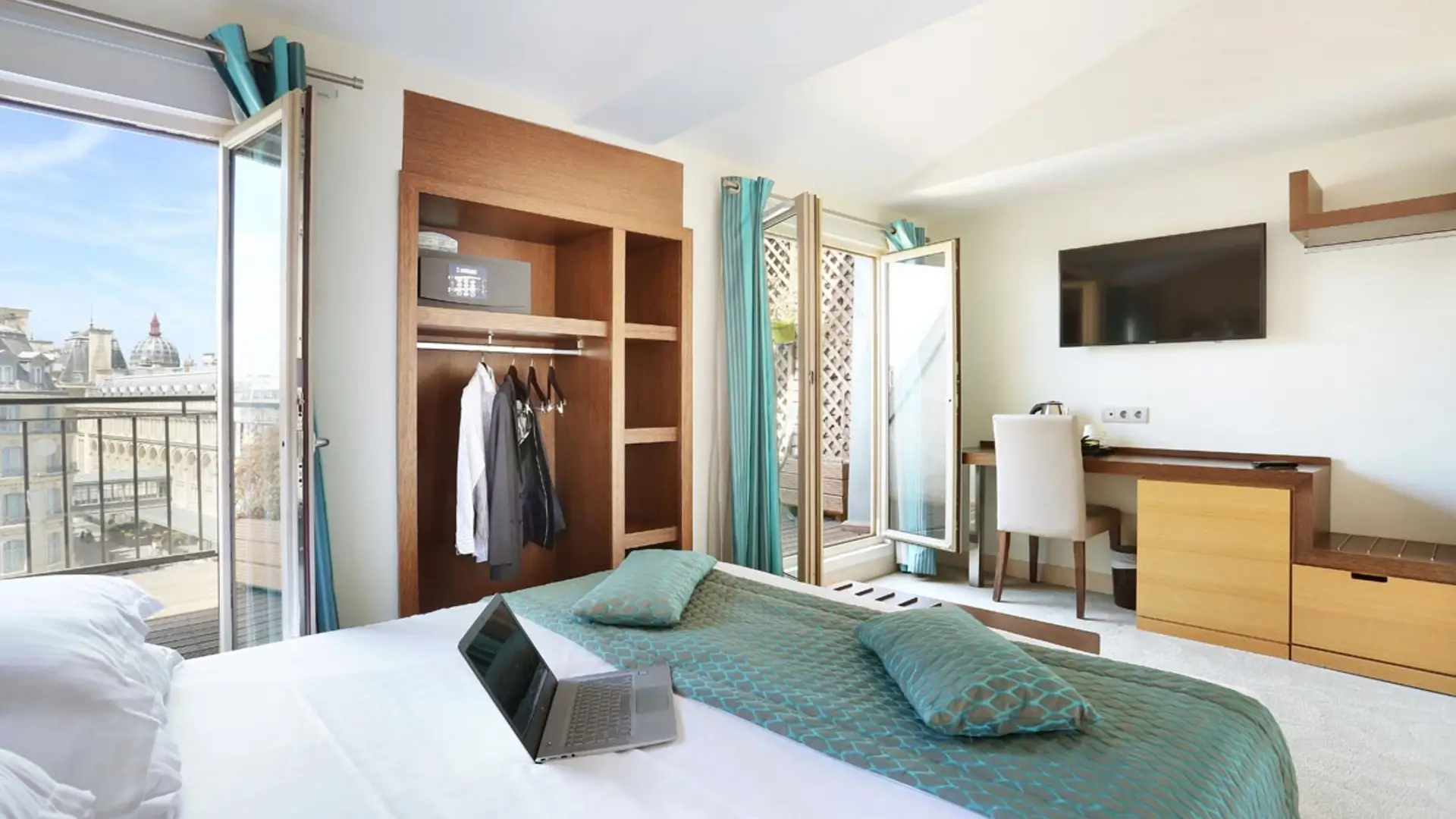 Hotel room with wardrobe, laptop on bed, and balcony over Paris rooftops.
