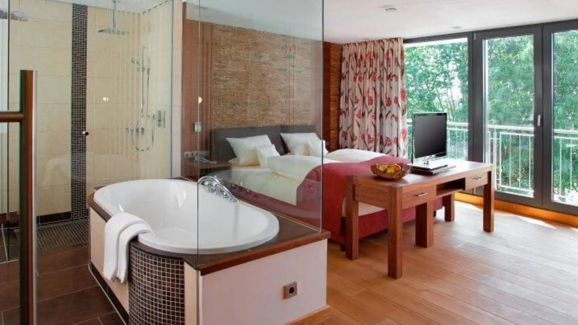 Open-plan room with glass-enclosed bathtub, bed, and forest view.