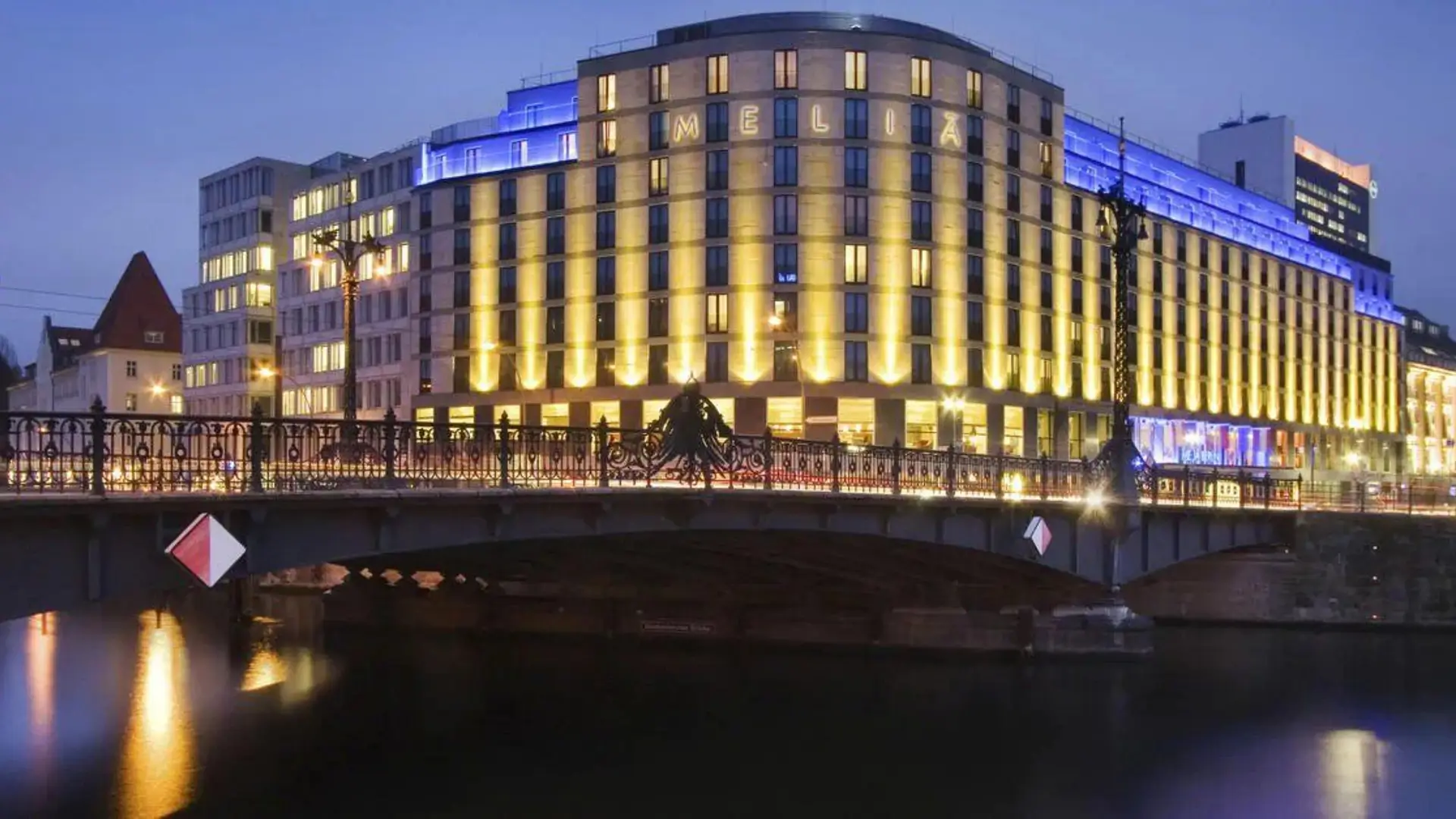Illuminated Meliá hotel at night, seen across a river bridge.