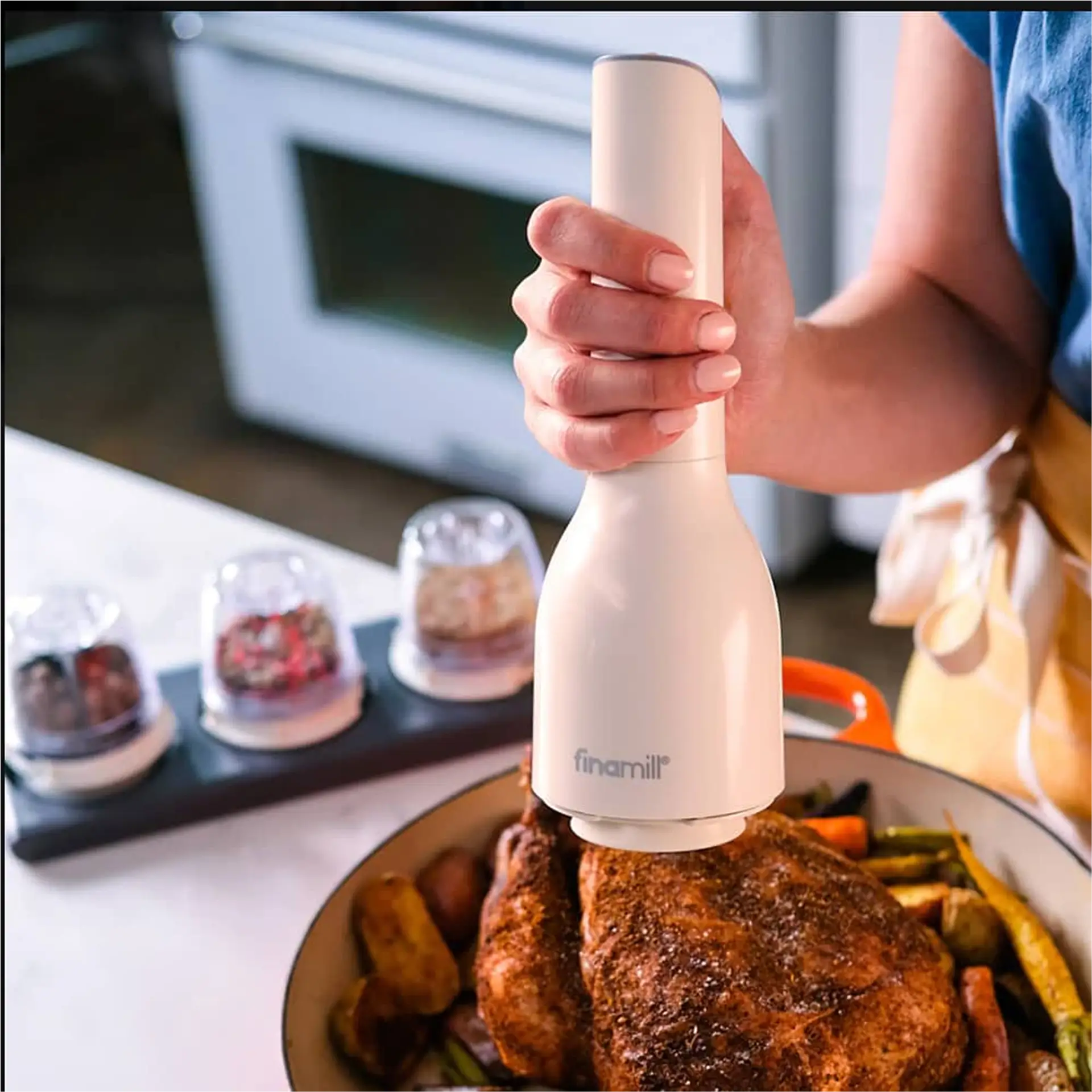 Hand holding white FinaMill grinder over roasted chicken and vegetables.