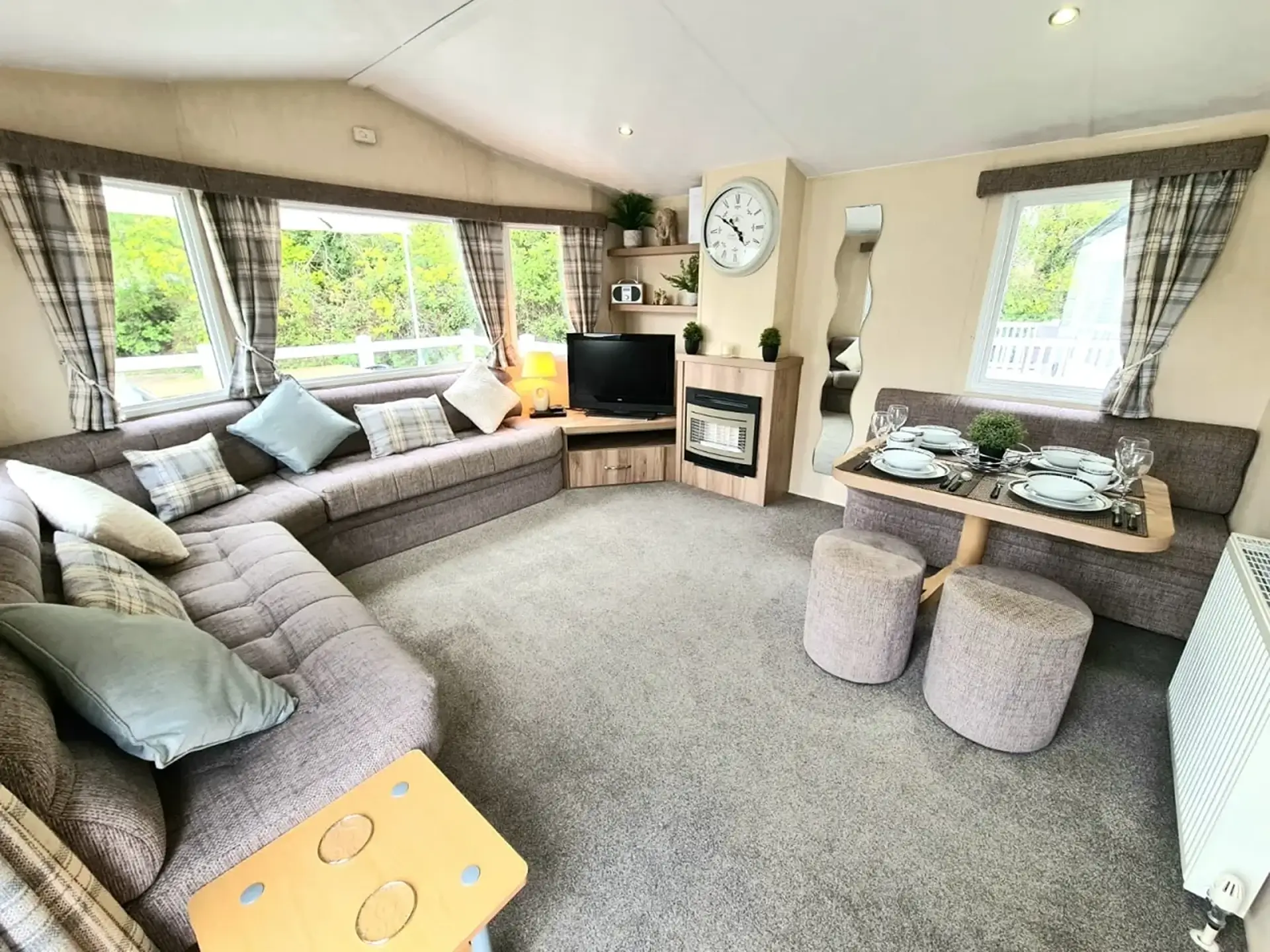 Corner sofa lounge with dining table, fireplace and clock, wooded view outside.