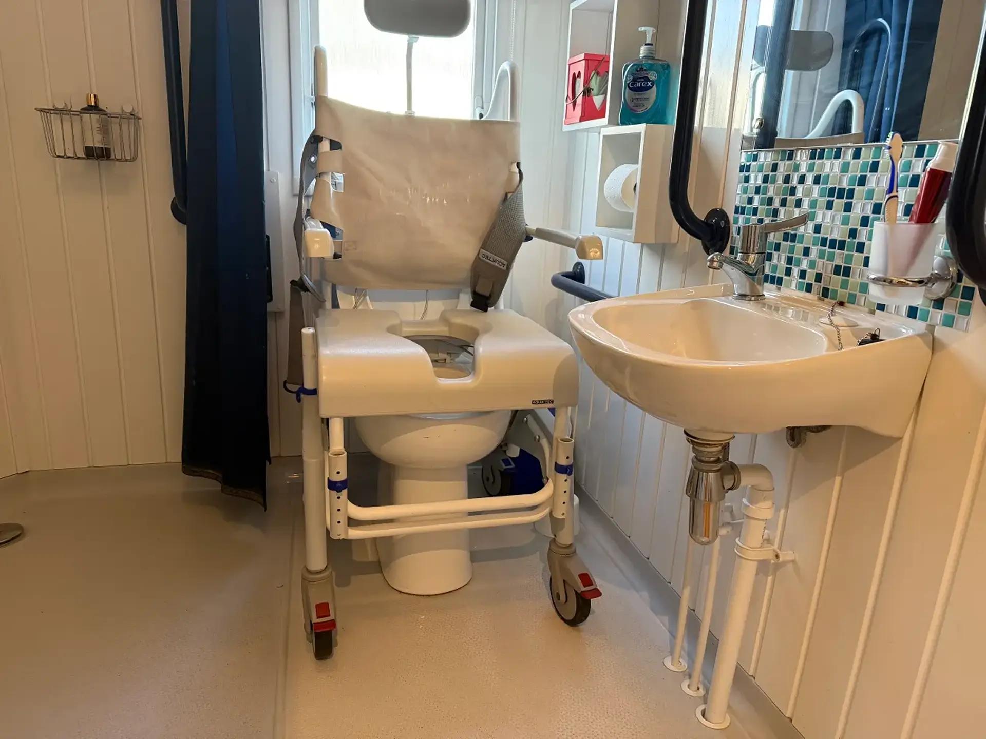 Wet room with a mobile shower/commode chair beside the toilet and sink.