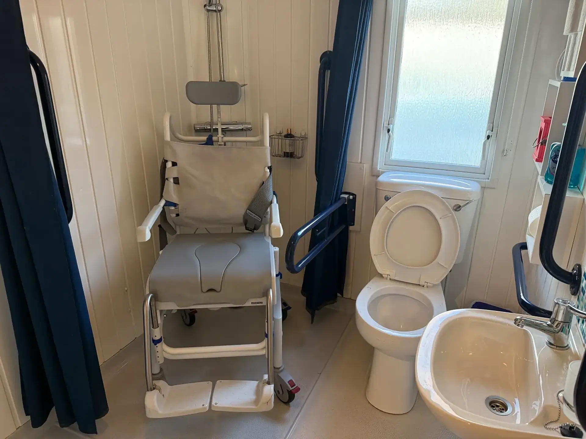 Wet room with a shower/commode chair beside the toilet, grab rail on the wall.