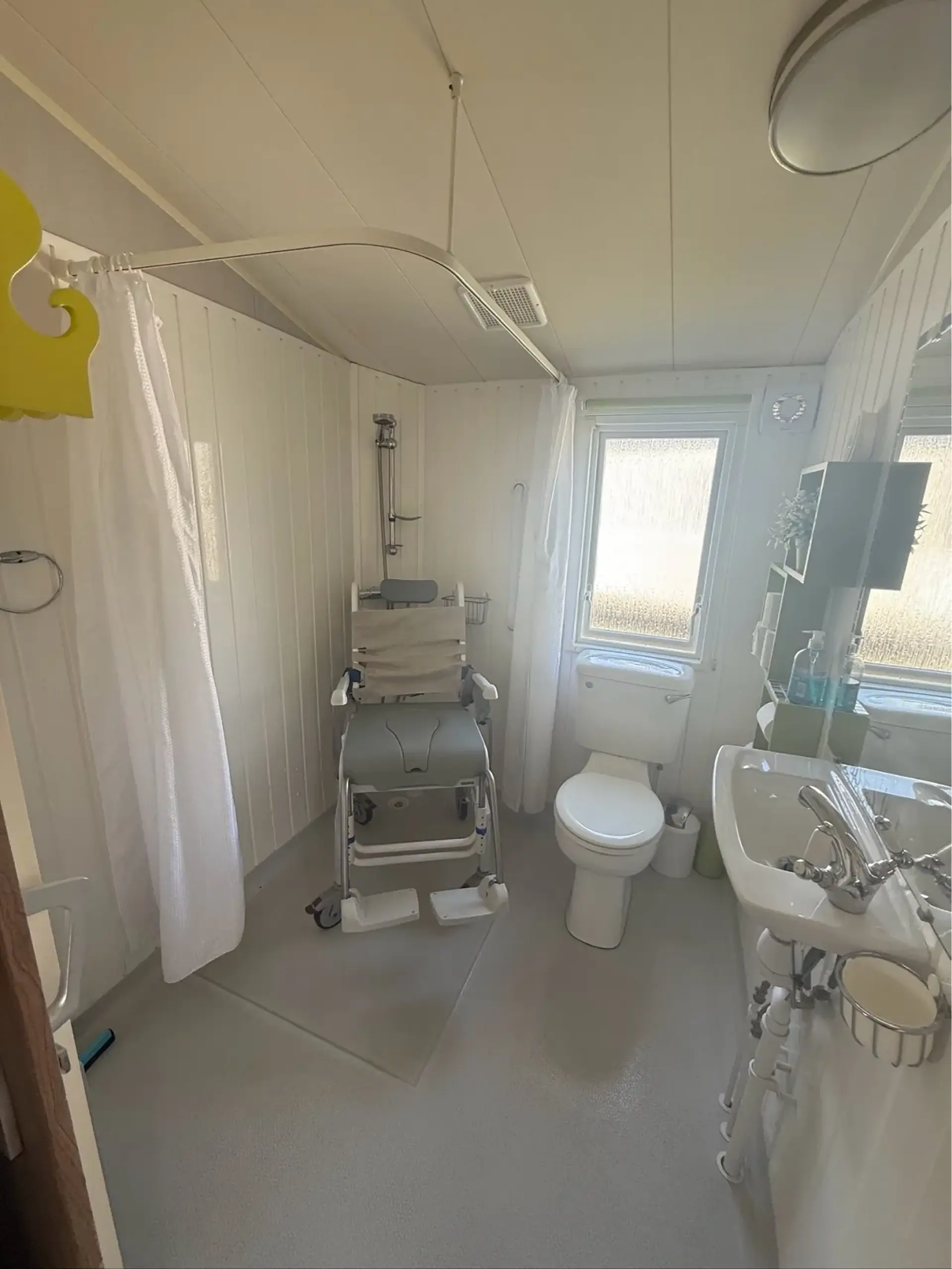 Wet room with shower chair, toilet and sink beneath a ceiling hoist rail.