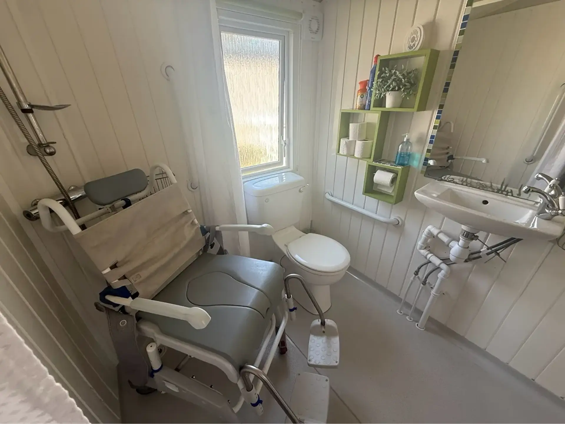 Wet room with a commode/shower chair, toilet, sink and grab rails.