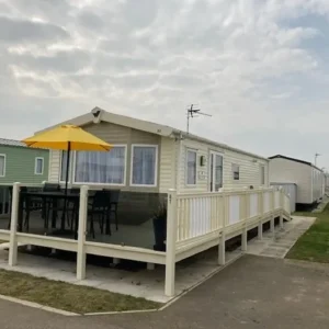 Cream holiday home with decking, yellow parasol and dining set, on a caravan park.