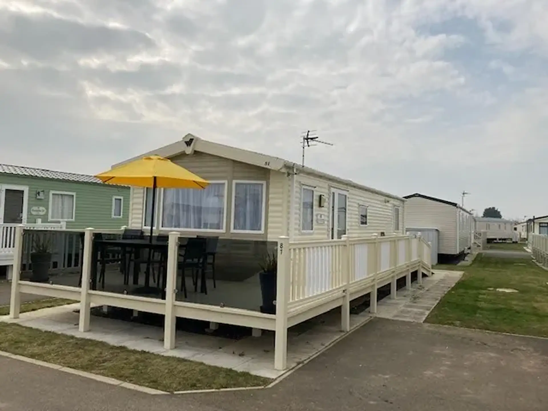 Cream holiday home with decking, yellow parasol and dining set, on a caravan park.