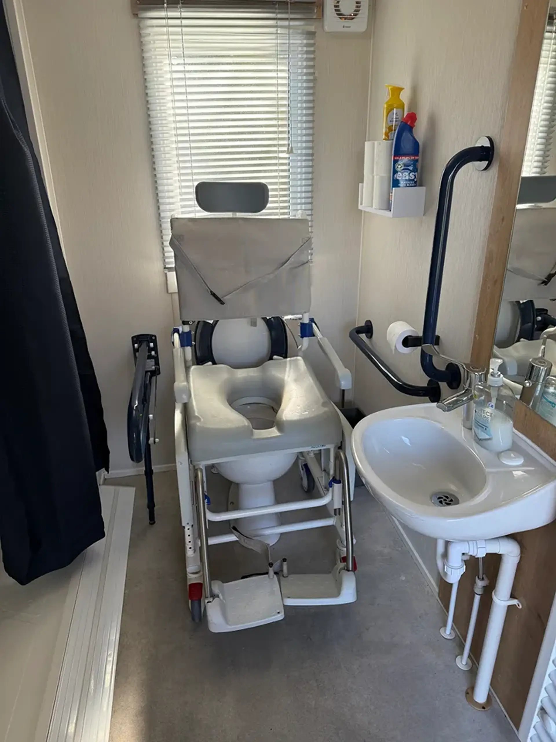 Bathroom with wheeled shower/commode chair, sink, and grab rail.