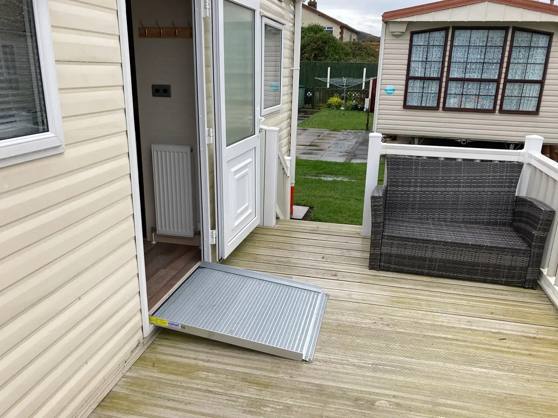 Portable ramp at caravan's open door leading onto decking with rattan sofa.