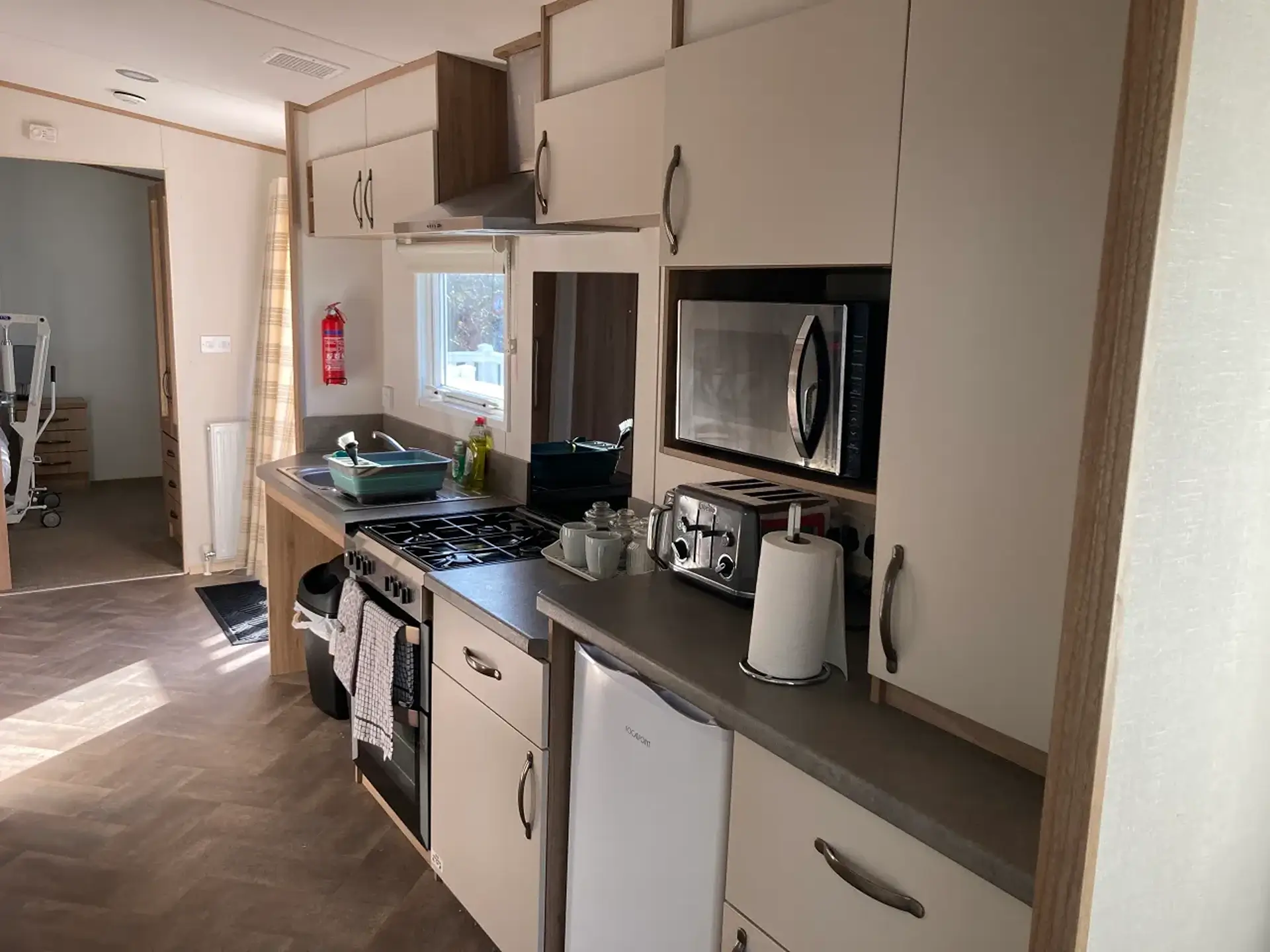 Galley kitchen with hob, oven, microwave, fridge, and mobility hoist visible in adjoining room.