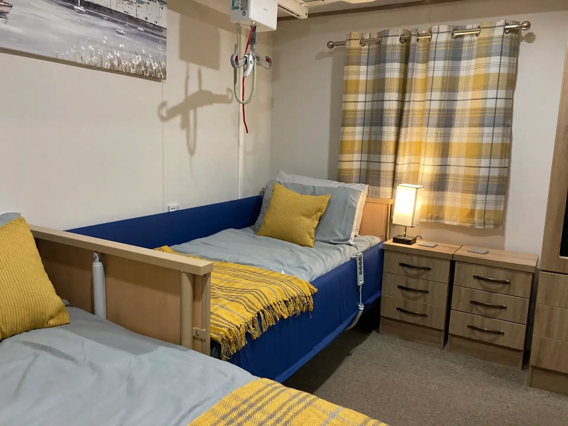 Bedroom with adjustable hospital-style bed, ceiling hoist track, and plaid curtains.