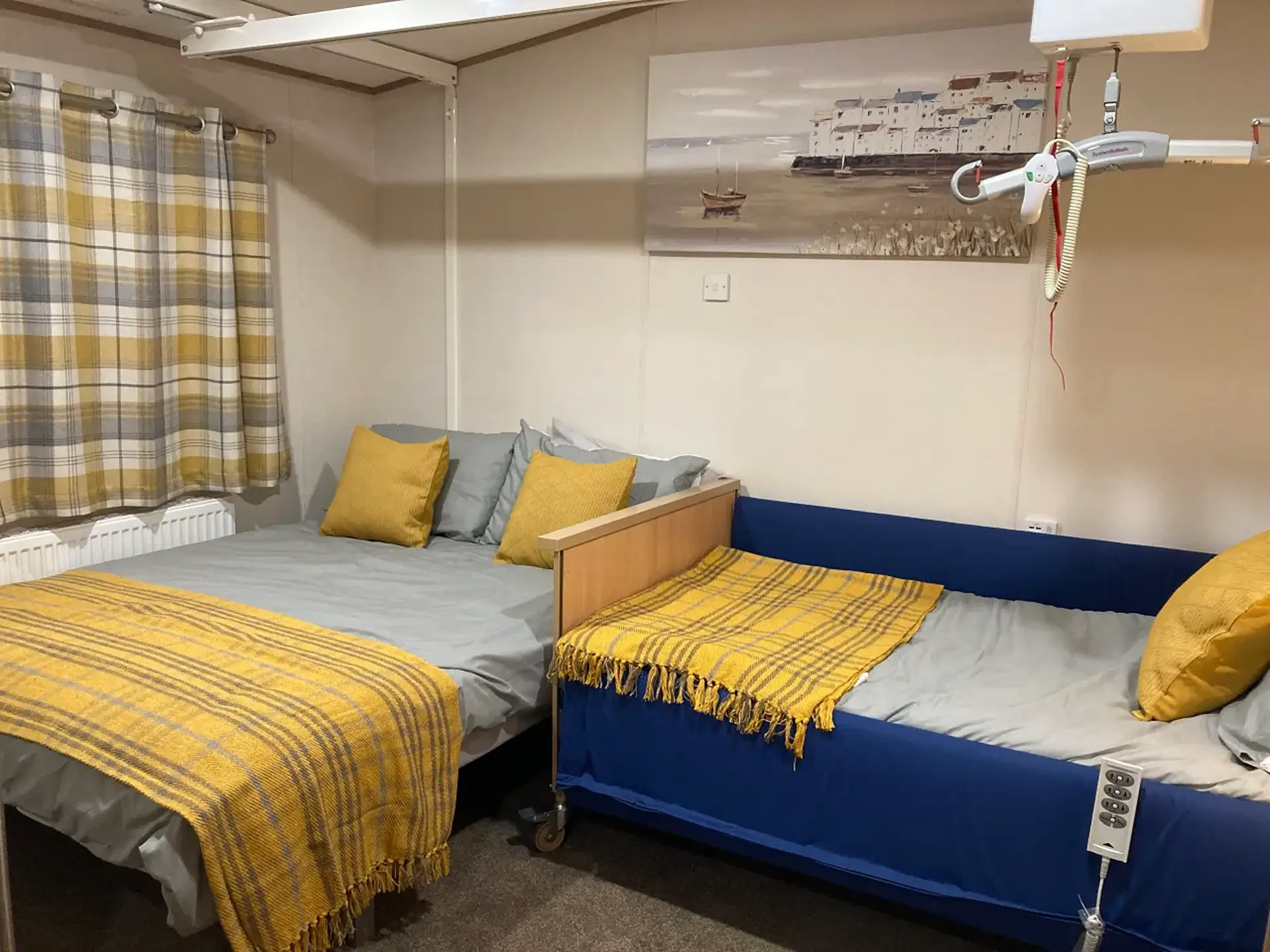 Two beds with a ceiling hoist track and coastal artwork above.