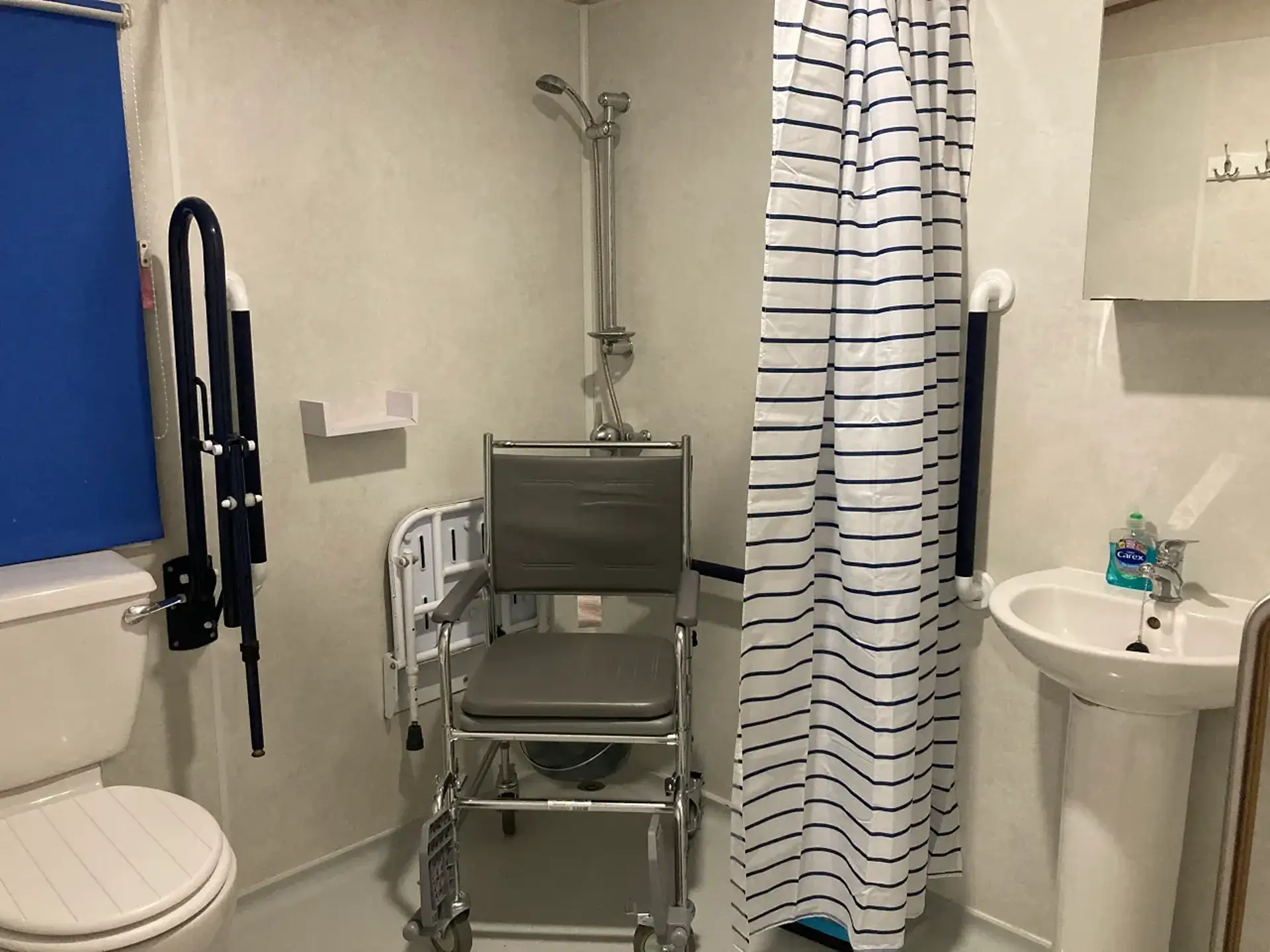 Wet room with wheeled shower/commode chair, grab rails, and striped shower curtain.