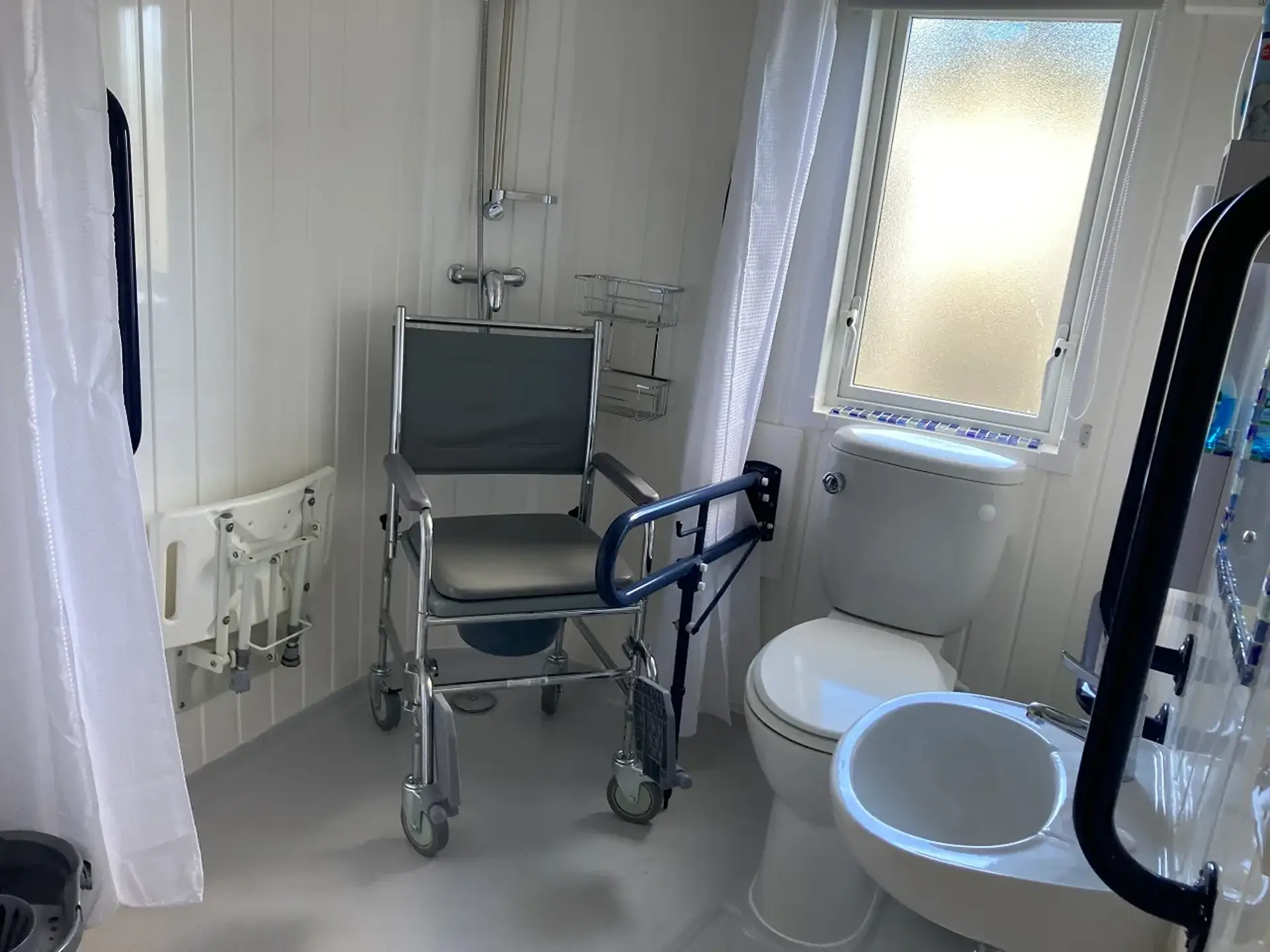 Wet room with wheeled shower/commode chair, grab rail, and toilet.