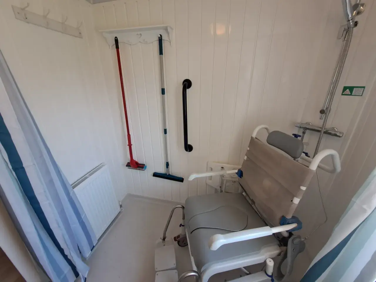 Wet room with shower chair, grab rail, and cleaning tools on wall.