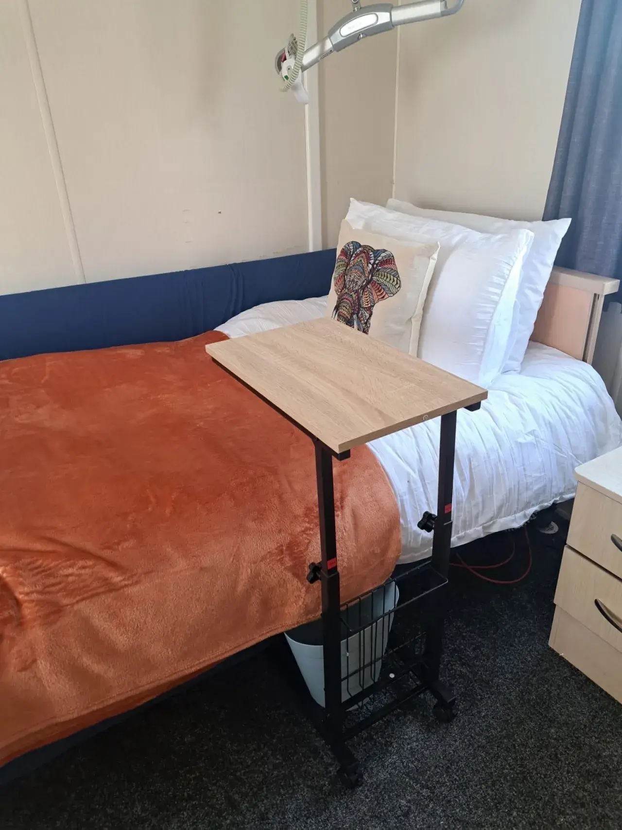 Adjustable bedside table over a bed with orange throw and cushion.