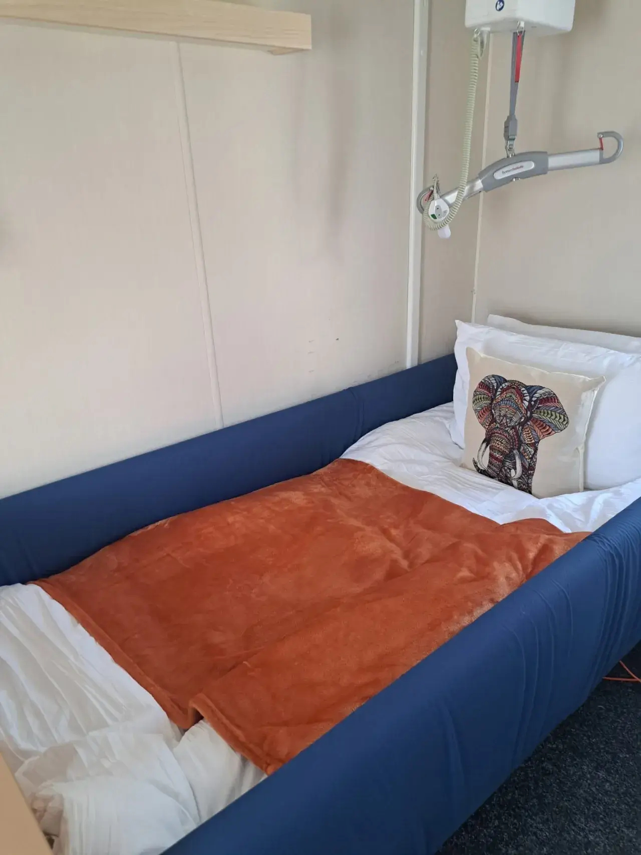 Close-up of a bed with blue side rails, orange throw, and ceiling hoist.