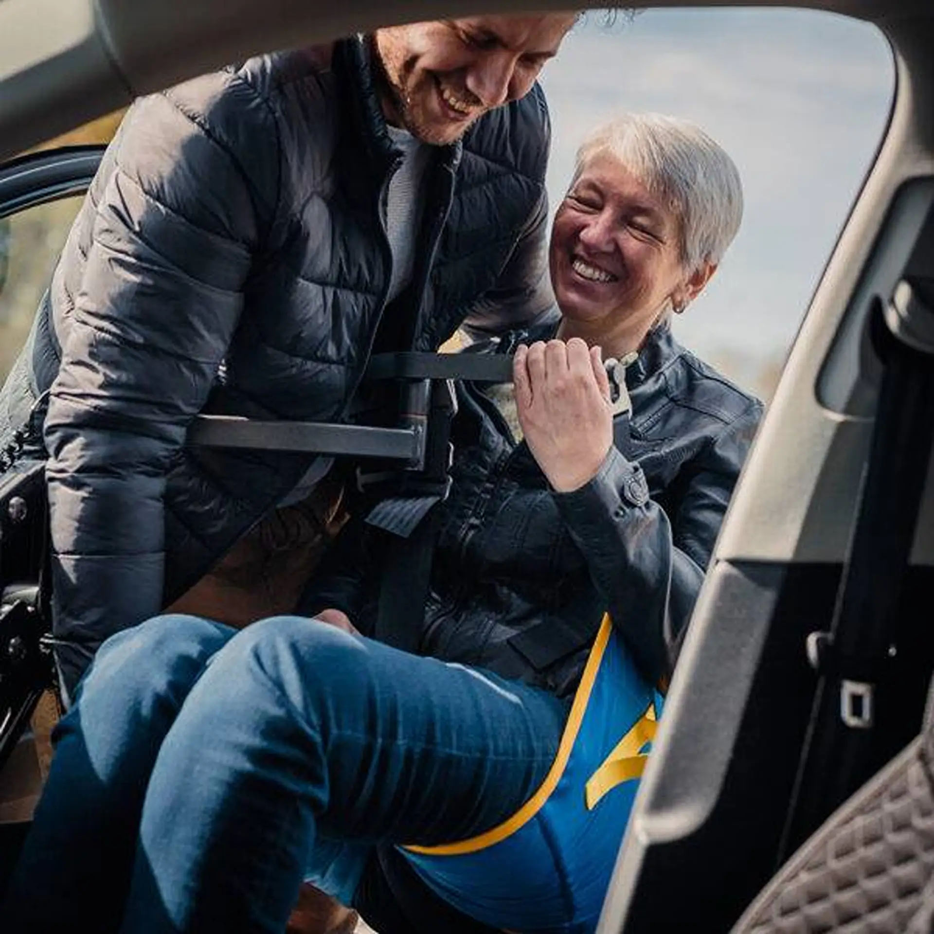 Man assisting a smiling woman being lowered into a car seat via the hoist sling.