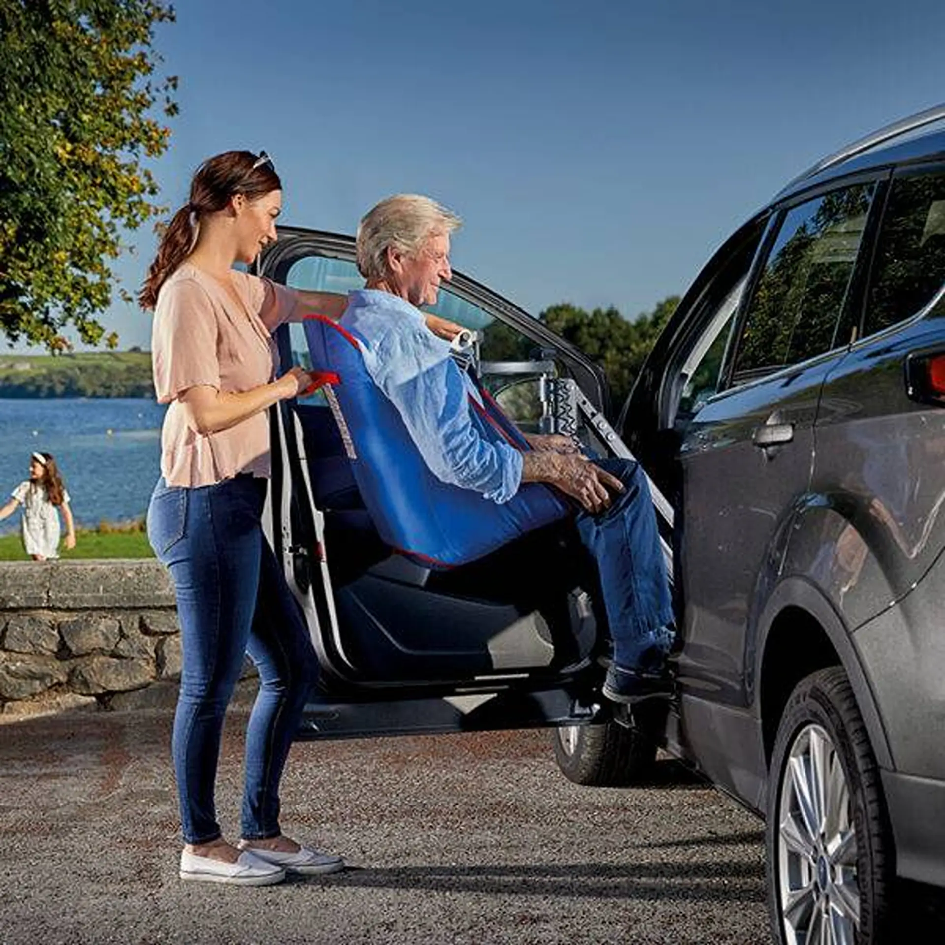 Woman assisting an older man being lowered into a car's passenger seat via the hoist.