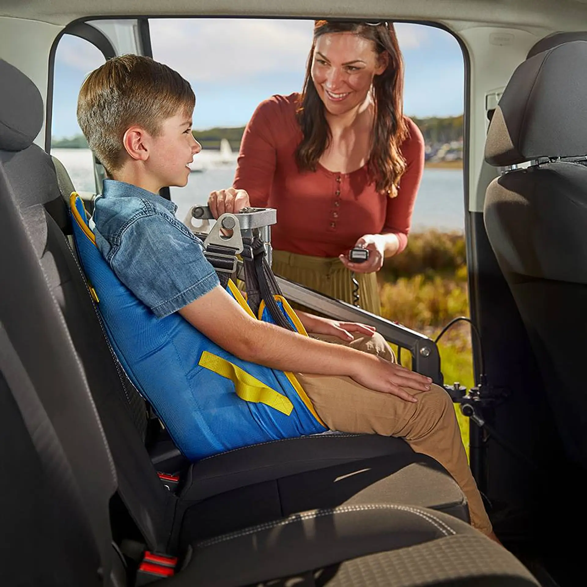 Boy in the hoist sling being lowered into a car's back seat by a smiling woman holding the remote.