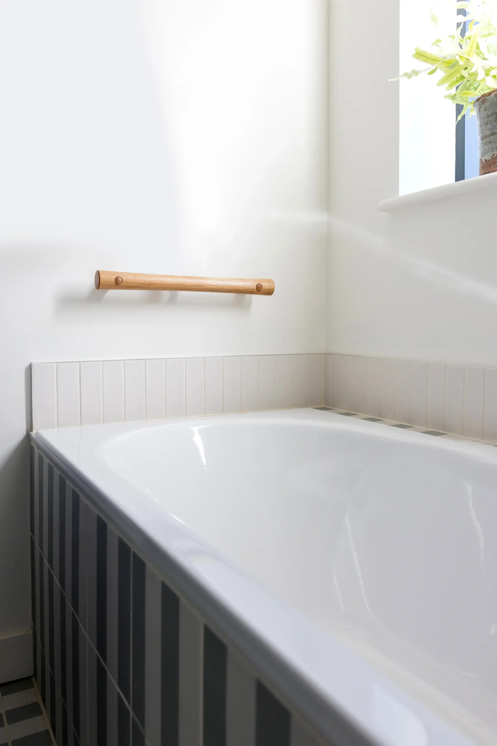 Straight oak grab rail mounted on wall above a bathtub corner, plant on windowsill.
