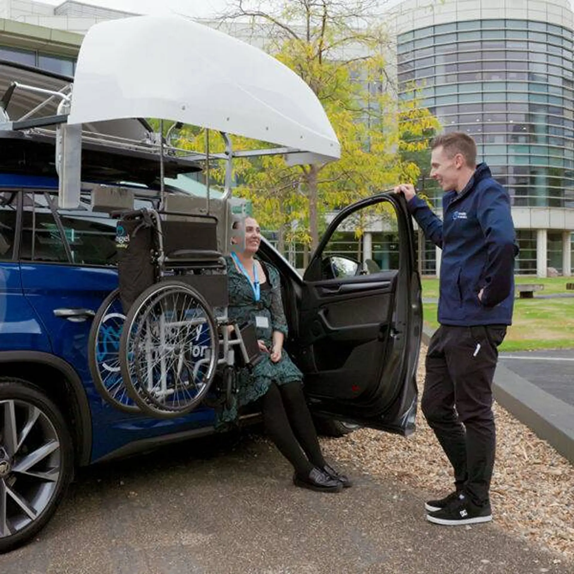 Woman in car doorway chatting with a man, wheelchair suspended from open rooftop topper