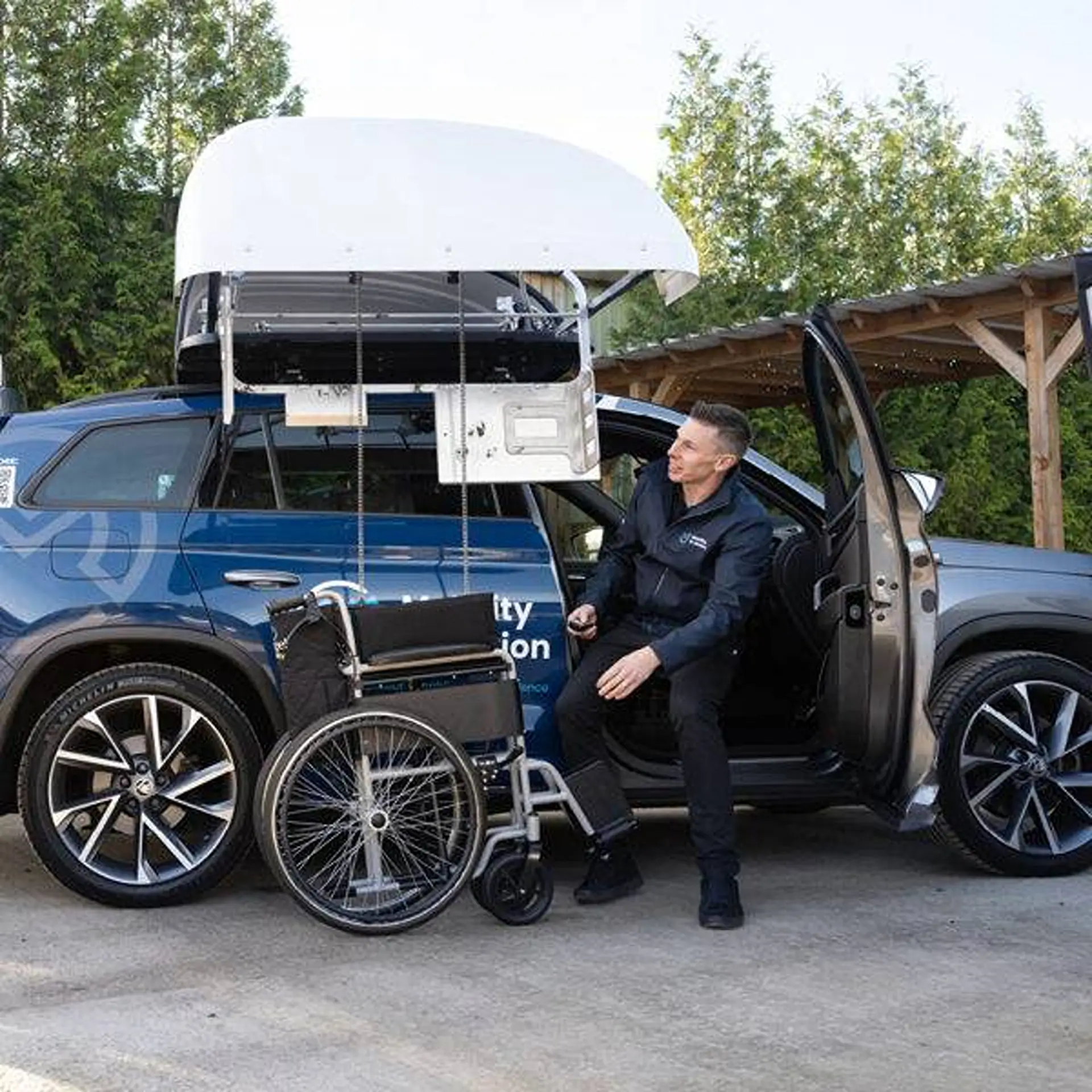 Man seated in SUV doorway, wheelchair lowered from open topper, remote in hand.