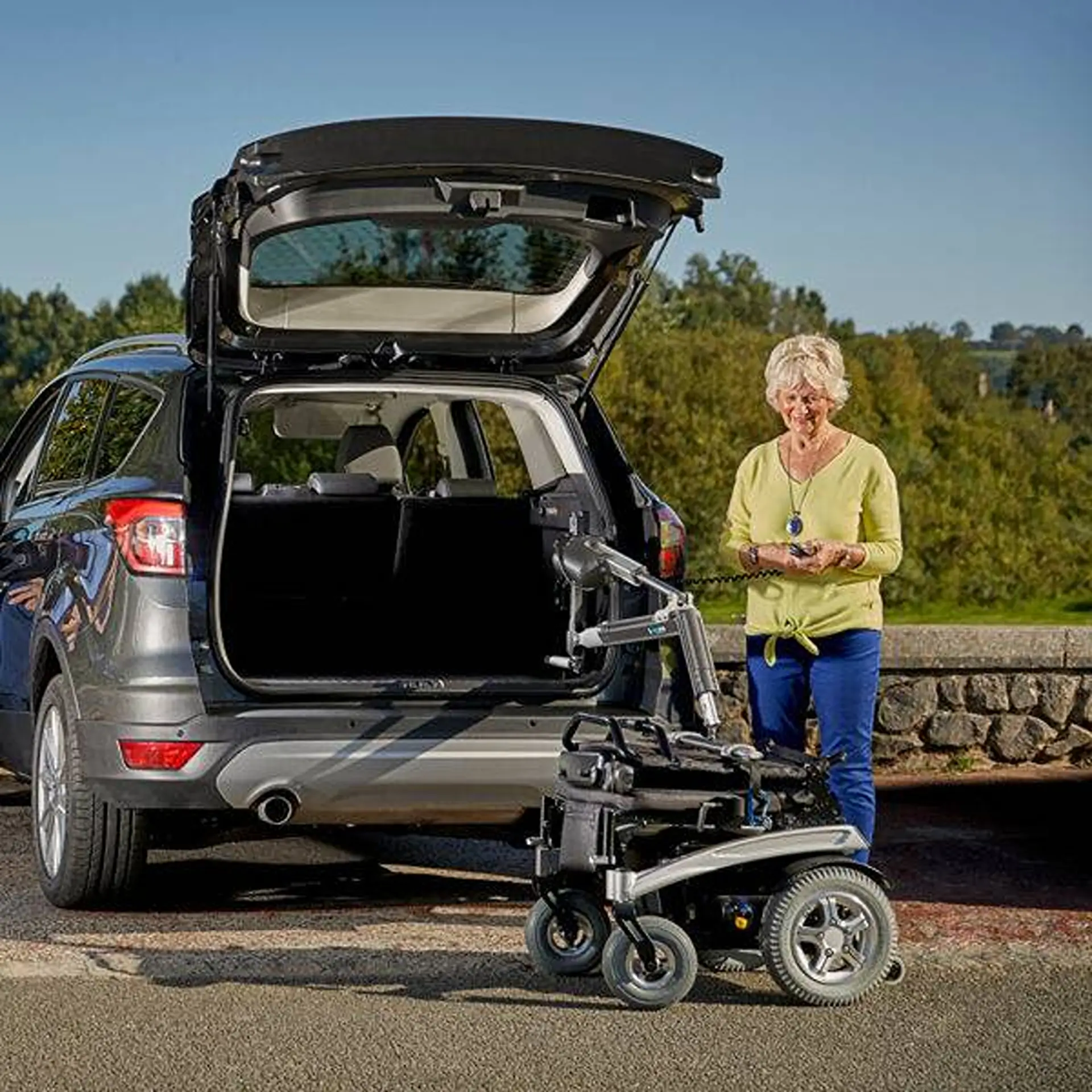 Elderly woman by a coastal car park operates the lifter arm holding a powered wheelchair outside her SUV's open boot.