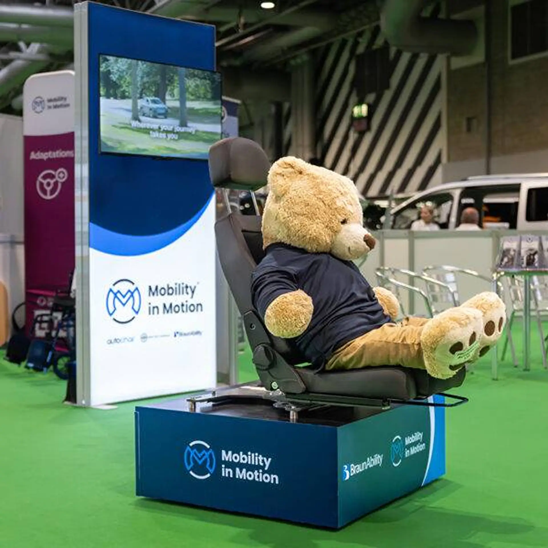 Teddy bear seated in a swivelled car seat at a trade show display stand.