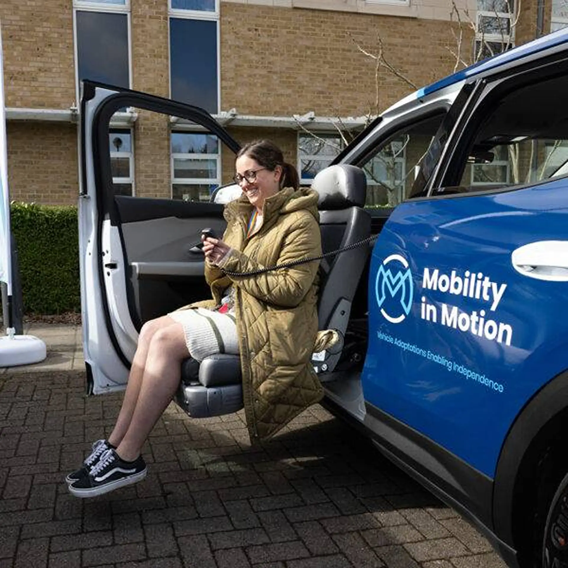Woman seated in a swivelled-out seat holding the remote, parked outside a building.
