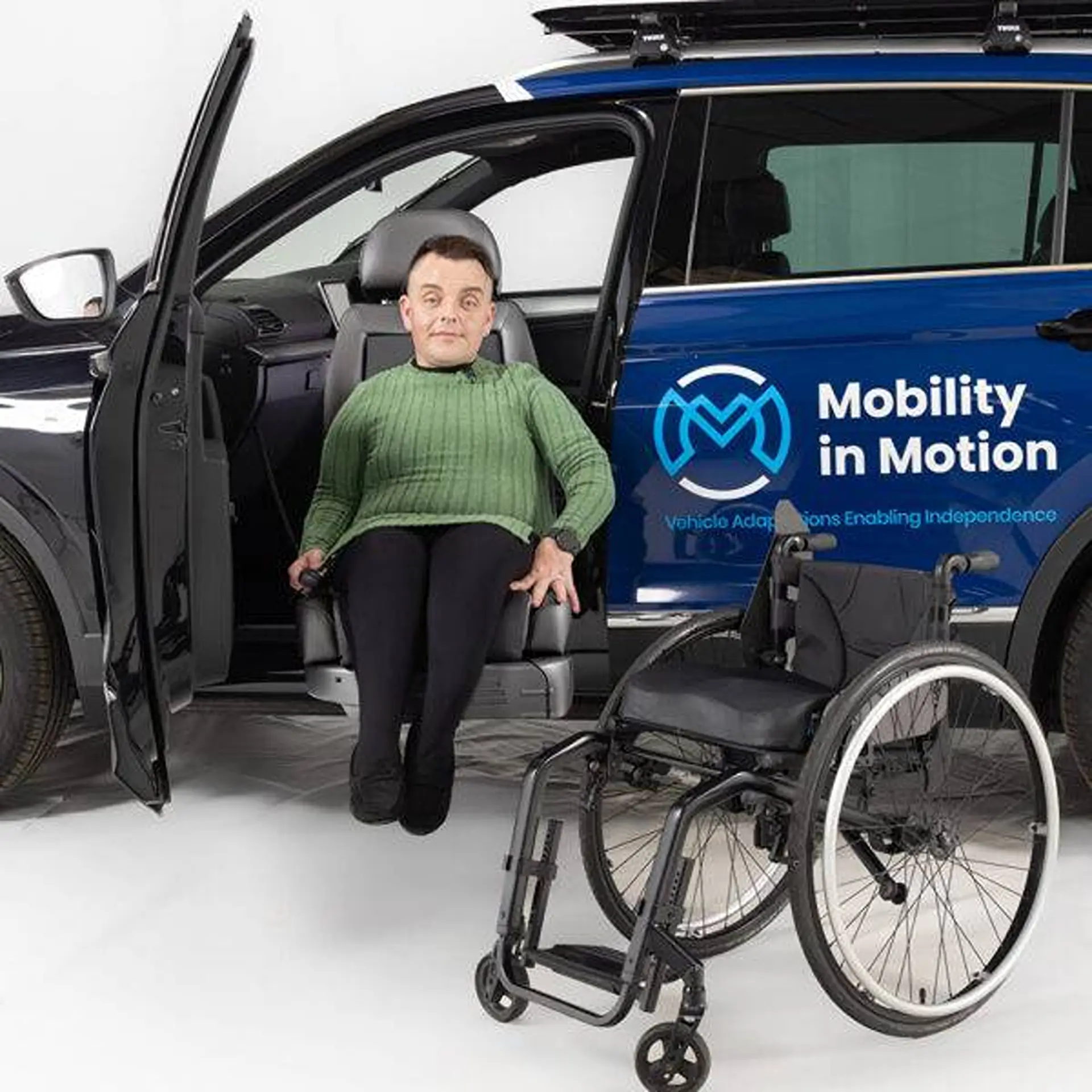 Woman seated in a swivelled-out car seat beside her wheelchair, studio background.