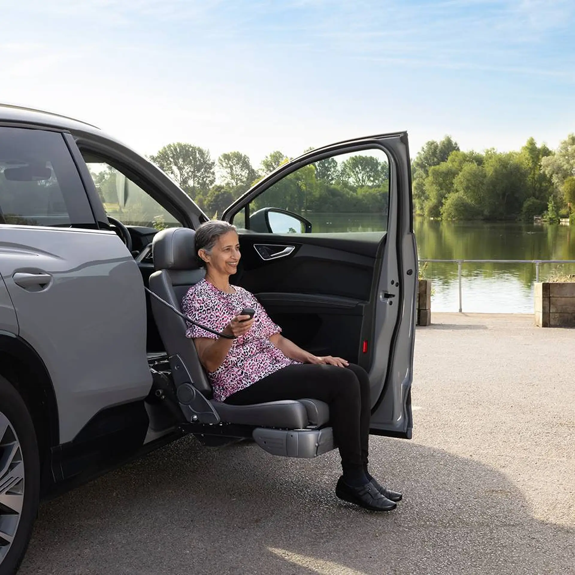 Woman seated in a swivelled-out car seat holding the remote, lakeside setting.
