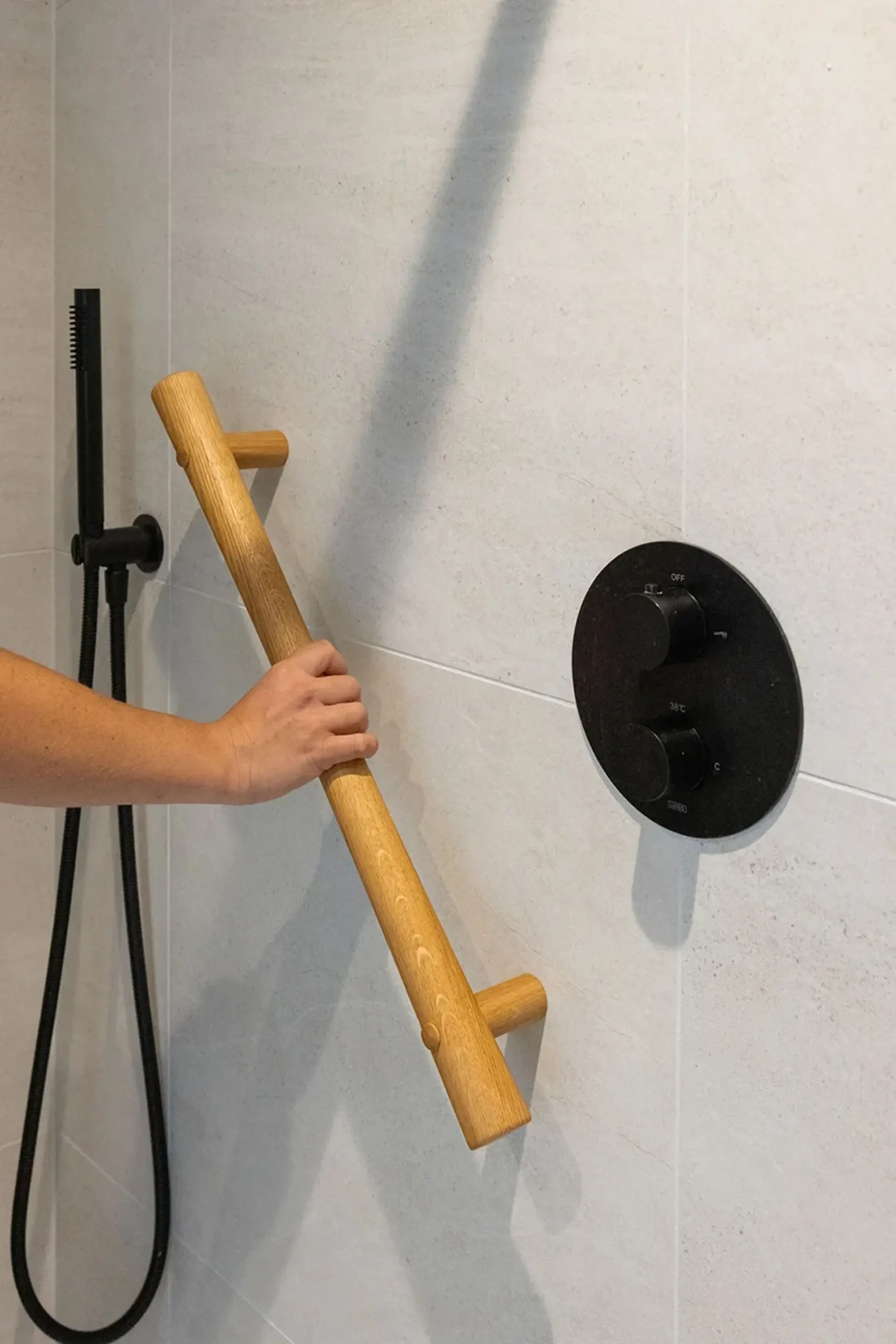 Hand gripping an oak grab rail in a shower near black controls.